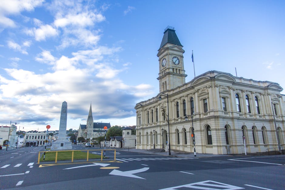 A wide-angle view of a historic municipal building featuring a tall clock tower with a peaked roof and decorative arched windows, situated on a city street. In the foreground, a paved area with marked parking bays and a small grassy island with a monument or obelisk surrounded by a low yellow barrier can be seen. To the left, a row of buildings and a church with a pointed spire are visible in the background. The sky above is partly cloudy with patches of blue, and the scene is well-lit with natural daylight. The setting appears to be a central area suitable for home relocation activities, with potential for loading and unloading furniture and packing materials, in line with professional removals services such as those offered by Man with Van Berrylands.