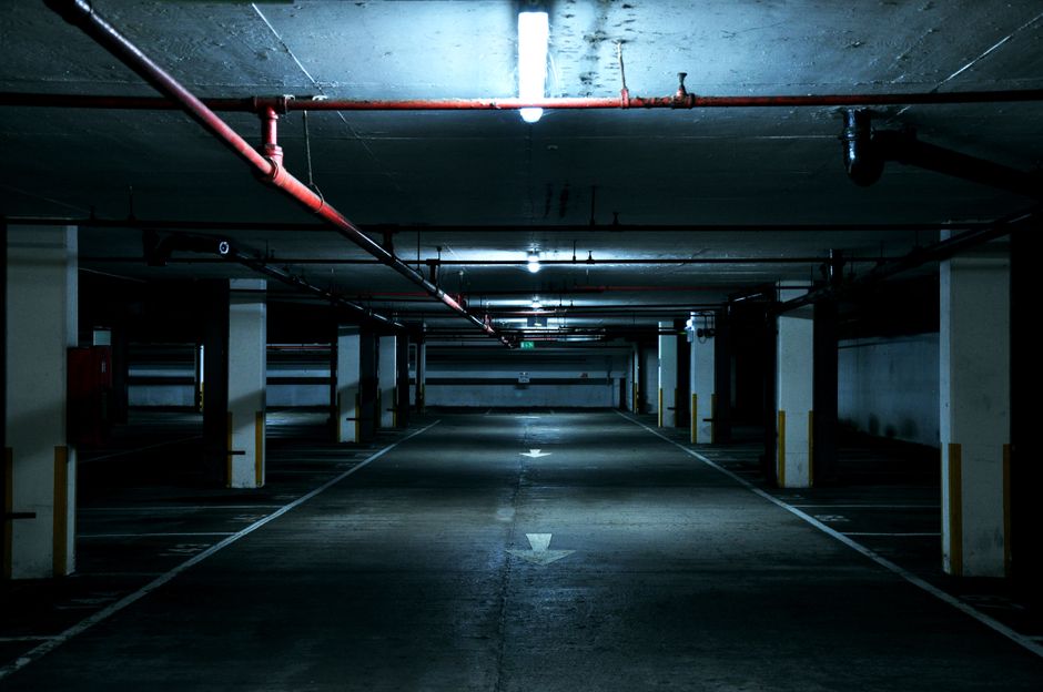 An empty underground parking garage with concrete flooring, supported by concrete pillars painted with yellow safety markings. Overhead, red and black pipes run along the ceiling, illuminated by fluorescent lights casting a cool, dim glow across the space. The parking bays are delineated with white lines, and some contain parked cars, though most are vacant. The central lane has white directional arrows painted on the ground to guide vehicle movement. The environment appears clean but somewhat industrial, with a focus on functional design suitable for vehicle parking and access for house removals and furniture transport. This setting reflects a typical location where a professional removal team, such as Man with Van Berrylands, might load or unload boxes and furniture during a house relocation, ensuring proper coordination and safety during the loading process.