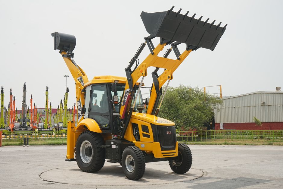 A yellow compact backhoe loader, with a front loader bucket and a backhoe arm, parked on a paved surface outdoors. The machine has a fully enclosed cabin with large windows, black tires with white rim