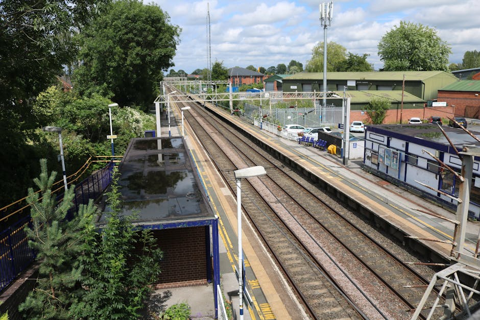 An elevated view of Berrylands train station platform showing the railway tracks running alongside. The platform is bordered by a blue metal fence and features a small brick building with a flat roof, which has puddles of water on top reflecting the cloudy sky. There is a lamppost near the edge of the platform, and several parking spaces with white and blue cars are visible in the background, adjacent to a green industrial warehouse with a sloped roof. Tall trees and green bushes border the station area, providing natural surroundings. Overhead electrical lines run parallel to the tracks, supported by tall metal poles. The scene captures the quiet environment typical of a suburban train station, suitable for home relocation logistics and furniture transport services by Man with Van Berrylands, emphasizing the space and infrastructure involved in moving and packing activities related to house removals and logistical planning.