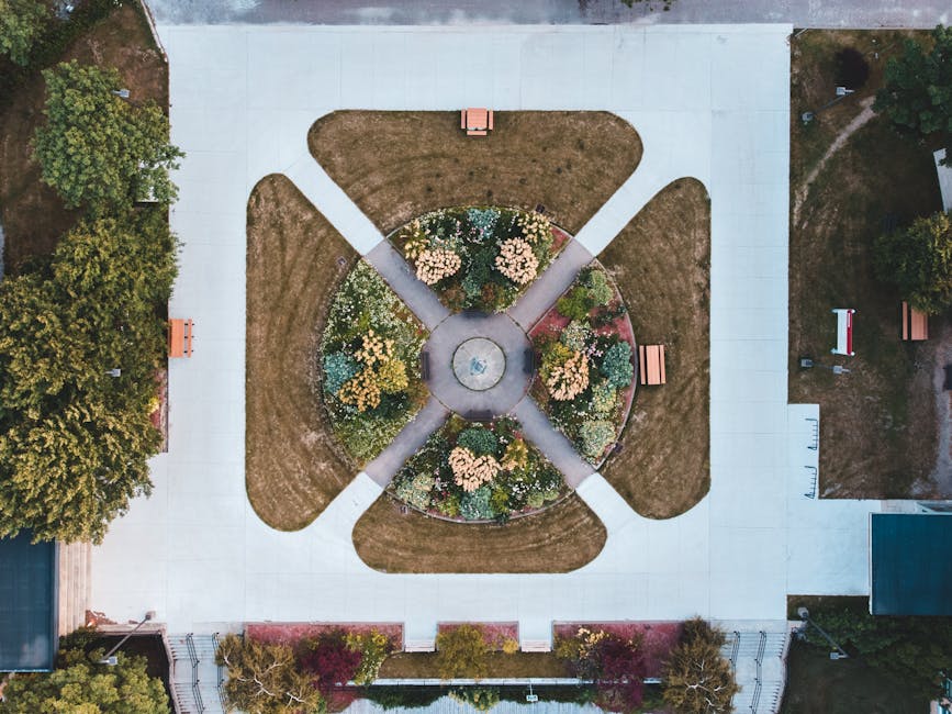 An aerial view of a circular flower bed with a central fountain, surrounded by four walking paths that extend outward in a cross pattern, located within a park area. The park features neatly mown grass sections, with several park benches situated along the pathways on a paved surface. To the left, there are dense trees with green foliage, and to the right, a small building or shed is visible. The scene is well-lit, with natural daylight illuminating the landscape. As part of a home relocation or furniture transport process, this image may depict the park area where Man with Van Berrylands is involved in careful planning for the logistics of moving large furniture or boxes to or from nearby properties, utilizing secure loading zones and ensuring smooth access for removal vehicles.
