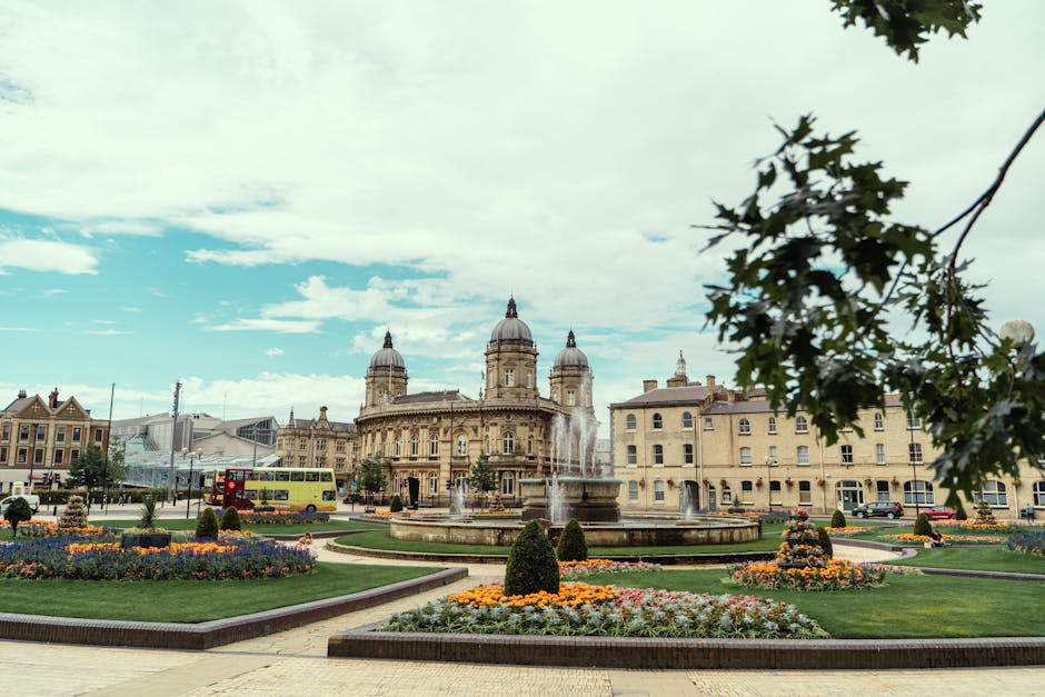 A wide view of a historic city square featuring a large, ornate stone building with multiple domes and decorative architectural details, set against a partly cloudy sky. In the foreground, there is a circular fountain with water jets, surrounded by well-maintained flower beds with colorful seasonal flowers, arranged in symmetrical patterns. To the left, a bright yellow double-decker bus is visible on the street, along with parked cars and pedestrians walking along the paved pathways. Some greenery, including a tree branch with green leaves, extends into the top right corner of the image, framing the scene. The environment is bright and clear, with natural daylight illuminating the textures of the building’s stonework and the vibrant colors of the flower arrangements. This setting suggests a lively urban area suitable for home relocation support, with identifiable transportation and public space elements. Man with Van Berrylands often operates in similar environments, assisting with packing and moving logistics in areas like Kingston Council with local permits for house removals.