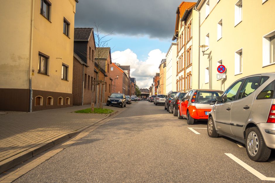 A narrow residential street in Berrylands with several parked cars along the curbside, including a silver van, a red hatchback, and other vehicles, with multi-storey buildings on both sides featuring a mix of brick and painted facades. The street is paved with asphalt and has a designated parking area marked by white lines. A no-parking sign is mounted on a wall on the right side of the street, indicating parking restrictions. In the background, there are various parked cars, a small tree with no leaves on the sidewalk, and a partly cloudy sky overhead. The scene is typical of an urban home relocation area, where vehicle parking and street access are relevant to moving logistics. Man with Van Berrylands often helps manage parking clashes during house removals, ensuring smooth furniture transport and loading processes in tight street environments.