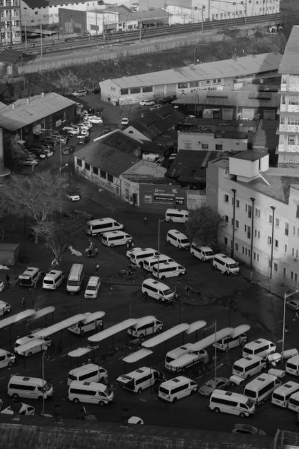 A black-and-white aerial view of a densely packed parking area on Berrylands streets, showing multiple white vans and small cars parked in various orientations across the asphalt surface. Some vehicles are parked parallel to the curb, while others are angled or irregularly positioned, indicating potential parking clashes or tight spaces. In the background, there are low-rise buildings and small commercial or residential structures with flat roofs and visible doors and windows. A few items such as cardboard boxes, plastic wrapping, and packaging materials are scattered around the open areas near the vehicles. The scene suggests ongoing home relocation or furniture transport activities, with vehicles possibly being loaded or unloaded in a busy urban setting managed by Man with Van Berrylands, a professional removals service specializing in city-based moving and packing logistics. The lighting is natural, with shadows cast by the vehicles and buildings, capturing the cluttered and active environment typical of a typical house removals operation in a congested street setting.