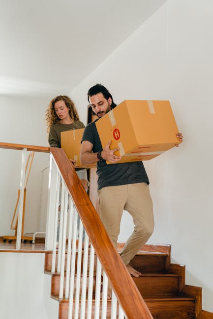 A man and a woman are carrying cardboard moving boxes on a staircase inside a residential property. The man, positioned at the front, is holding a medium-sized box with both hands, wearing beige trousers and a dark top, focused on the task of home relocation near a wooden staircase with white banisters. The woman, following behind, is also holding a cardboard box, dressed in casual clothing, and walking down the stairs with her attention directed towards the task. The interior environment features white walls and a ceiling, with natural lighting illuminating the area. In the background, part of a hallway and additional boxes can be seen, indicating the process of packing and furniture transport as part of a home moving operation. This scene reflects the loading process involved in professional removals services, such as those provided by Man with Van Berrylands, during a same-day emergency move in Berrylands.