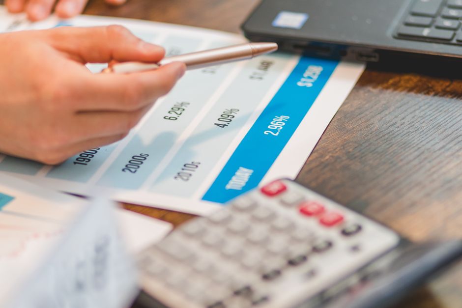 Close-up view of a person's hand holding a silver pen over a financial chart printed on a white sheet of paper, which displays numerical data in columns and a blue-highlighted section. The paper is pl