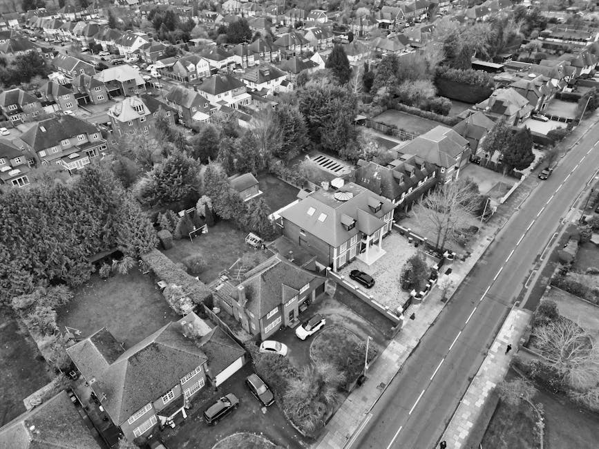 An aerial black-and-white photograph of a residential area showing narrow streets lined with closely spaced houses and buildings, with numerous parked cars along the pavement and in driveways. The sce
