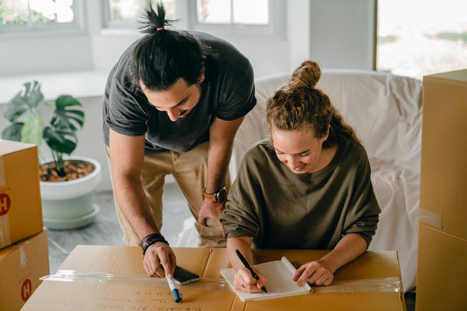 A man and a woman are engaged in house removals inside a well-lit room with large windows, preparing for a home relocation. The man, wearing a dark t-shirt, is leaning over a large cardboard box on a table, holding a marker and a smartphone, possibly planning or checking a packing list. The woman, dressed in an olive-green top, is seated at the same table, writing on a notepad with a pen. Surrounding them are several packed cardboard boxes sealed with clear packing tape, some with printed labels and others with handwritten notes, indicating organized packing for a furniture transport or moving process. In the background, there is a large white sofa, and a potted plant with broad leaves adds greenery to the space. The scene depicts the detailed steps involved in packing belongings for a house move, coordinated by a removal company such as Man with Van Berrylands, emphasizing efficient packing and the logistics of unloading and transporting household items.